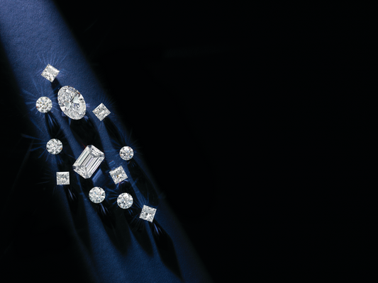 Assorted diamond cuts including round, oval, emerald, and princess shapes displayed under a spotlight on a dark background